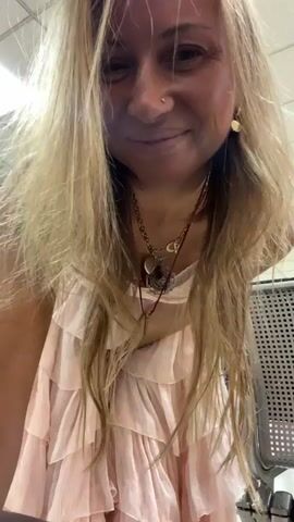 A woman with blonde hair wearing a pink lace bralette and a pink tiered ruffled skirt spins in a circular motion. The video is taken from a low angle, showing the woman's face looking up and smiling. She is in an indoor environment with fluorescent lighting and what appears to be an airport waiting area with seating and vending machines in the background.