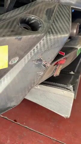 The video shows close-up shots of a green and yellow race car with carbon fiber body panels. The first shot shows damage to a section of the car near the exhaust pipe, with a piece broken off. A red tag is visible nearby. The next shot shows a Pirelli sticker on the car. Then, the camera focuses on a Tm Performance logo on the side of the car near the rear wheel. Finally, the video shows the side of the car, including the tire and the "GRUPPO FENERA" writing on the side. The car is parked on a red floor.