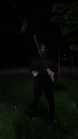 A person wearing a black hooded sweatshirt and pants and a black mask with eye holes holds two katanas on a lawn at night. The swords are held out to either side, then one is brought to the front. A wooden fence and a tree are in the background.