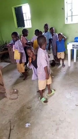 A group of children are gathered in a room. The room has green walls and a concrete floor. There are windows with bars on them and a table with a sign that reads "UBEC SUBEB 2022 INTERVENTION." The children appear to be playing a game, with one child in the foreground wearing a plaid shirt and shorts, holding a white object. Other children are standing around, watching and participating. The overall atmosphere seems lively and playful.