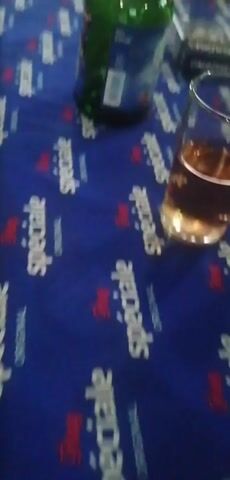 The video shows a table covered with a blue tablecloth featuring a repeating pattern of text and logos. On the table are several items, including a glass containing a amber-colored liquid, two lighters (one red and one green), an ashtray with cigarette butts, a bottle with a cork, a pack of cigarettes, and a green bottle. The lighting is dim, suggesting an indoor setting, possibly a bar or a home. The camera pans across the table, focusing on the various objects.
