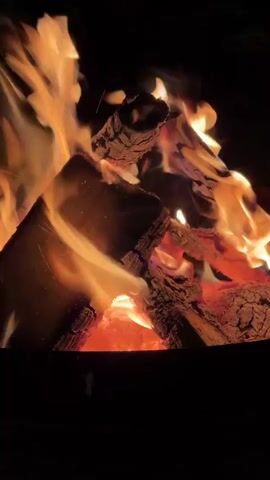 The video shows a close-up of a campfire at night. Logs are burning, with flames and glowing embers visible. The background is dark. No people are visible. The focus is on the fire.