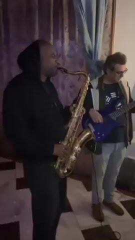 The video shows three men performing music in a cluttered room. The first man, wearing a black hoodie, plays the saxophone. Next to him, a second man with glasses plays a blue bass guitar. The camera pans to a messy table filled with electronic equipment, wires, a laptop, and a DJ mixer. A man wearing a red beanie and headphones is seen working at the DJ mixer, adjusting controls. The room has patterned floors, curtains, and a ladder leaning against the wall. There are stacks of records and a lamp on a shelf.