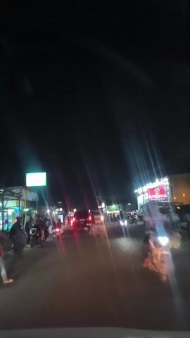 The video shows a nighttime scene on a busy street. Several cars and motorcycles are visible, some with their headlights on, illuminating the road and surrounding area. People are gathered on the sidewalks and near businesses. Bright lights from storefronts and streetlights create a vibrant, somewhat chaotic atmosphere. The camera appears to be inside a moving vehicle, suggesting the video was captured from a car or similar mode of transport. The overall impression is of a lively commercial area at night, with traffic and pedestrian activity.