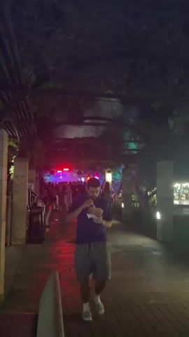 A man walks in the foreground eating from a container. Behind him, a pathway is lit by colorful lights. People are visible in the background, and there are buildings and trees. The scene appears to be an outdoor public space at night.