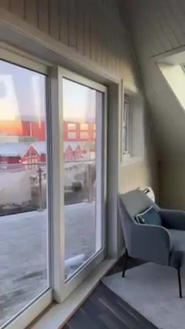 The video shows the interior of a room with large windows offering views of a waterfront scene. The room appears to be an attic or top-floor space, characterized by sloping ceilings and white wood paneling. There is a large sliding glass door that opens to a wooden deck covered with snow. Outside the door, red buildings are visible in the distance. The scene outside the windows includes snow-covered rocks, water, and a distant shoreline with what appears to be mountains. Inside, two matching gray armchairs are placed on a patterned rug. A small table sits next to one of the chairs, holding decorative items like candles and a plant. The room is well-lit, with natural light streaming through the windows, suggesting it is either sunrise or sunset. A voice can be heard in the background.