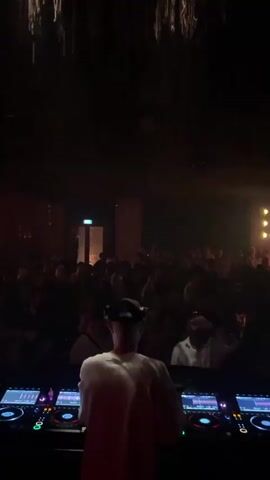 The video opens from a first-person perspective, looking over a DJ's setup at a crowded nightclub. The DJ, wearing a white t-shirt and a baseball cap, is facing a crowd of people. The club is dimly lit with red and yellow lights, and there is haze or smoke in the air. The DJ's equipment, including turntables and a mixer, is in the foreground. The crowd is densely packed, with most people appearing to be dancing or socializing. As the video progresses, the lighting in the club changes, with more yellow lights becoming prominent. The camera remains focused on the DJ's perspective, with the crowd filling the background. There are no clear interactions between the DJ and the crowd, but the overall atmosphere suggests a lively party or event. The video ends with the yellow lighting intensifying.