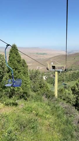 This is a vertical, handheld video shot from a ski lift moving upward. The ski lift is a bright yellow pole with a mechanism at the top, from which hangs a single ski lift chair in the foreground. Another ski lift chair, colored blue, is visible on the left side of the frame, slightly further away. Several more ski lift chairs, some carrying people, can be seen traveling up the cable behind the yellow pole. The ski lift is surrounded by lush green trees and vegetation, with a rolling, green landscape stretching out into the distance under a clear blue sky. In the background, a valley is visible, with a hazy, distant city or town at the horizon. The overall impression is a scenic mountain view on a sunny day.