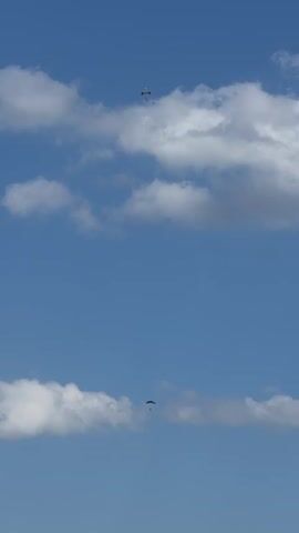 Two individuals are skydiving against a bright blue sky with scattered white clouds. The parachutes are visible, one higher than the other. Both skydivers appear to be in descent. The clouds are fluffy and white, indicating fair weather. The scene is captured from a low angle, looking up at the sky.