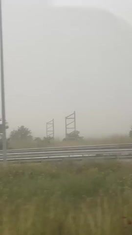 The video shows a view from inside a moving vehicle, looking out at a foggy landscape. The road is lined with a concrete barrier and a metal guardrail. There are trees and grassy areas visible on the other side of the road. Streetlights and a large billboard structure occasionally pass by. The fog is thick, obscuring much of the view and giving the scene a muted, overcast appearance.