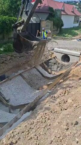 In Hîncești, Moldova, an excavator is depositing concrete into a trench. Two construction workers, dressed in high-visibility vests and hard hats, oversee the operation. The trench has wooden forms in place to shape the concrete. A concrete culvert with a circular opening is visible nearby. The scene appears to be an outdoor construction site, possibly for infrastructure development or road repair. The time of day seems to be daytime, and the weather appears clear. The overall atmosphere is one of active construction work.
