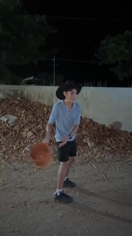 At night, in an outdoor area with dirt ground, a person wearing a light blue polo shirt, dark shorts, dark shoes, and a dark cowboy hat throws a large brown pot towards a wall. The pot breaks when it hits the ground. The person then jumps and throws their hands up while looking forward. In the background is a wall, some trees, and a pile of broken brown pots. There is an electric light on a pole.