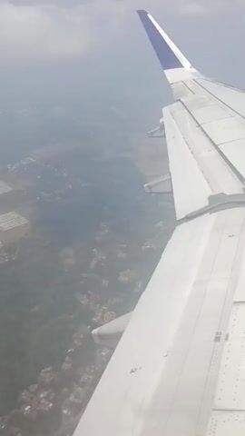 The video shows a view from an airplane window, looking down at the ground below. The wing of the plane is visible in the foreground, and the landscape is hazy, suggesting it is either cloudy or polluted. The ground appears to be a mix of rural and suburban areas, with some buildings and trees visible. There are no people or animals visible in the video. The video is shaky, indicating that the plane may be in motion or experiencing turbulence.
