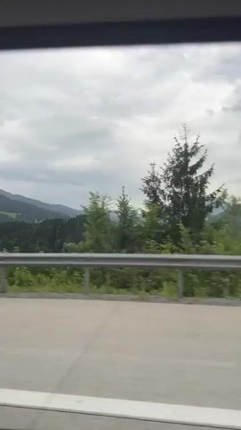 A car is driving on a highway. The passenger side window view shows a cloudy sky, mountains in the distance, green trees and vegetation, and a metal guardrail. The highway pavement is visible in the foreground with white lane markings.