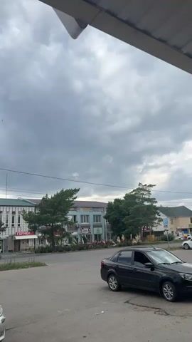 This media file captures an overcast day in Yegorlykskaya, Russia. The scene is a street view, with a road in the foreground and buildings lining the background. A black sedan is parked on the right side of the road. The sky is filled with heavy, dark clouds, suggesting an impending storm or a recent one. A few birds are visible in flight against the cloudy sky.

The buildings appear to be a mix of commercial establishments. On the left, a building with a white facade is decorated with a repeating geometric pattern. One sign on this building reads "ГЕРМЕС". Next to it, a building with a blue facade features several windows and what appears to be air conditioning units. Another sign on this building is partially obscured. Further down the street, more buildings are visible, some with green roofs.

A lamppost stands prominently in the center-right of the frame. The overall mood is somewhat somber due to the dark, cloudy weather, but the presence of the birds hints at life continuing despite the atmospheric conditions. The time of day is difficult to ascertain precisely due to the cloud cover, but it appears to be daytime. The location cues, such as the Cyrillic text on the signs, confirm the setting as Russia.