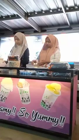 Two women are captured working behind a food stall counter in Sekopek Wetan, Indonesia. Both are wearing hijabs; the woman on the left wears a light grey/cream hijab and a dark top, while the woman on the right wears a light brown/tan hijab and a lighter-colored top. They are actively engaged in preparing or arranging items on the black countertop.

The front of the counter features a vibrant banner with a pink and purple gradient background. It showcases four illustrations of clear cups filled with layered drinks, likely bubble tea or dessert beverages, featuring jelly, whipped cream, and fruit toppings. Prominently displayed on the banner is the text "So Creamy!! So Yummy!!" in large, stylized white letters.

On the counter, various items are visible: the woman on the left is near a woven basket containing small containers or bottles, and a white container with a green lid. The woman on the right is handling small items in her hands, with stacks of black plastic containers nearby. In the background, a white sign or menu board shows partial text, including "ORIGINAL" and "ING CREAM."

The stall is covered by a corrugated metal roof, suggesting an outdoor or semi-outdoor setting. The lighting is bright and even, indicating either daytime or a well-lit evening. The scene depicts a casual, small-scale food business.