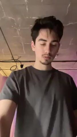 A young man with dark hair and a trimmed beard stands in a room with a rough concrete ceiling. He is wearing a gray t-shirt. Visible wiring and a light fixture hang from the ceiling. In the background, there is a hint of pink and blue light. The man is looking down and appears to be inspecting something, then looks briefly to his left. He then looks down again. There is an indistinct white noise in the background.