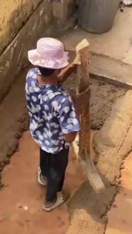 A person in a blue and white floral shirt, dark pants, sandals, and a pink hat is working on a wet cement floor. They are using a wooden tool with a horizontal bar to press and smooth the cement. There is an old concrete wall behind them, and a bucket, level, and other items are in the background. The person appears to be tamping down the wet cement.