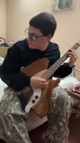 A young man with glasses is sitting on a bed and playing an electric guitar. The guitar's body appears to be modified or possibly built from a broom or a similar bristled object. He is wearing a black hooded sweatshirt and camouflage patterned pants. The room is simple, with a bed, a side table, and a picture on the wall. The lighting is soft and even. The man is focused on playing the guitar, looking at the fretboard and strings as he strums.