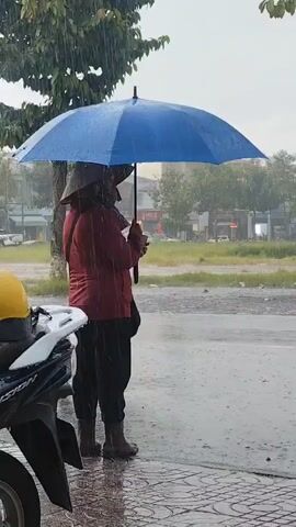 A person stands under a blue umbrella in the rain. They are wearing a conical hat and a red jacket. A motorbike is parked to the left of the frame. A car drives by in the background. The ground is wet and reflects the light.
