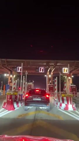 A video taken from within a moving car shows a dark gray Toyota SUV passing through a toll booth at night. The booth is brightly lit with overhead lights and has red and white striped barriers. The vehicle has a Moroccan license plate, which is illuminated by the car’s taillights. The driver is moving slowly towards the exit, where the barrier arm is raised and a green light signals that the vehicle is free to pass. Once through the toll gate, the SUV merges onto a highway lined with streetlights. The night sky is very dark.
