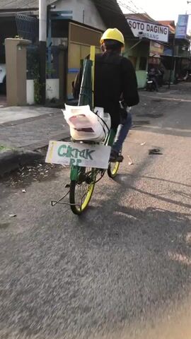 The video shows a man riding a bicycle down a street. He is wearing a yellow hard hat and a black jacket. The bicycle is green with yellow wheels. On the back of the bicycle, there are several items strapped on, including a plastic bag filled with white objects, some green poles, and a sign that says "Ciktak PP". A bottle of water is attached to the handlebars. The man is riding on an asphalt road, and there are buildings and a motorcycle visible in the background. The lighting suggests it is either early morning or late afternoon.