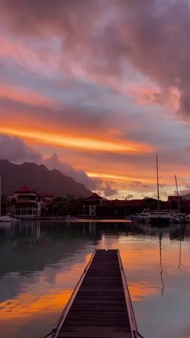 The video displays a picturesque harbor at sunset. In the foreground, a wooden pier extends into calm water, reflecting the vibrant colors of the sky. The water itself is a deep blue-green, with subtle ripples disturbing its surface. On the left side of the frame, several houses with red roofs are nestled amongst lush greenery and trees, with mountains visible in the distance. On the right, a cluster of sailboats and yachts are docked, their masts piercing the sky. The sky above is a dramatic spectacle of color, with fiery oranges, soft pinks, and deep purples blending together. Clouds are scattered across the sky, adding texture and depth to the sunset. The overall atmosphere is serene and tranquil, capturing the beauty of a coastal evening.