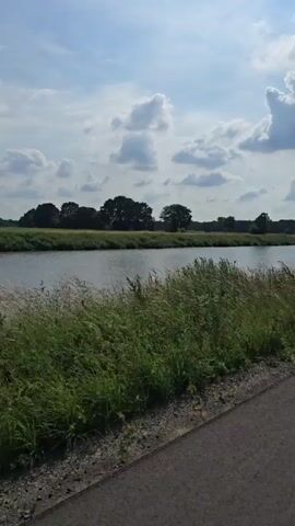 A young person on a bicycle rides down a paved path next to a canal. The path is bordered by tall grass and a canal with trees and fields on the opposite bank. The sky is partly cloudy and the sun is shining. The person is wearing a helmet and a white shirt. They are riding away from the camera. The video is a recording from a drone.