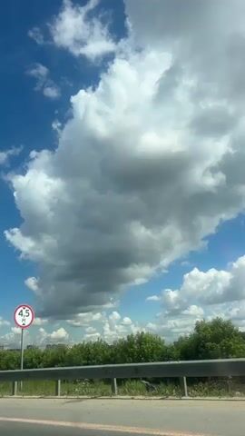 This is a vertical video taken from a moving vehicle, looking upwards and outwards towards a bright blue sky filled with large, fluffy white clouds. The camera pans slightly to the left. In the foreground, a grey asphalt road is visible, bordered by a metal guardrail. Beyond the guardrail, there is lush green vegetation, including trees and bushes. A traffic sign with a red circle and the number 4.5 inside it, indicating a height restriction, is visible on the right side of the frame. Further in the background, some low buildings and more trees can be seen. The overall impression is a sunny day with dynamic cloud formations.