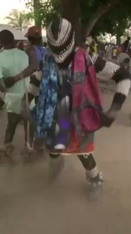 The video shows a person dressed in a colorful costume and mask dancing in a dusty outdoor area with other people around. The dancer wears a black and white knit mask, a colorful patchwork shirt, and what appear to be ankle bells. The ground is dusty, and there are trees and buildings visible in the background, indicating a village setting. People are seen moving around the dancer, some watching, some dancing, and some clapping.  A hand wearing a silver bracelet is visible briefly in the foreground. The scene appears to be part of a cultural celebration or performance.