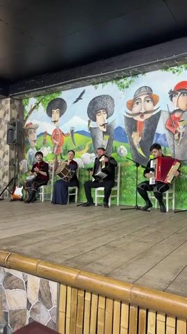 Five musicians are performing on a wooden stage in front of a mural. From left to right, the first musician is playing a drum. The second musician is also playing a drum. The third musician is singing into a microphone and playing an accordion. The fourth musician is playing an accordion. The fifth musician is playing a guitar. The musicians are all wearing traditional clothing. The mural behind them depicts several men with large mustaches and hats, as well as mountains and trees. The stage is decorated with bamboo.