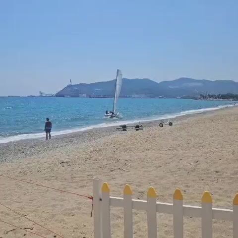 A wide shot of a sandy beach with a person standing near the water's edge. A small sailboat with two people on board is sailing in the turquoise water, close to the shore. There are two sets of boat trailers on the sand. In the background, there are mountains and industrial structures. The sky is clear and blue.