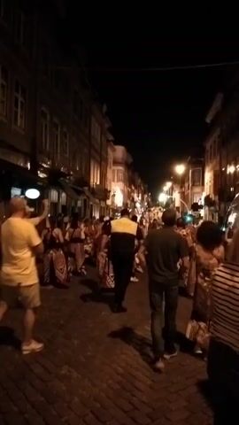 A group of people are walking down a cobblestone street at night. The street is lined with buildings, some of which have lights on. There are streetlights illuminating the scene. Some people are dressed casually, while others appear to be dressed for an event. One person in the center of the frame is wearing a yellow shirt and beige shorts. Another person in the foreground is wearing a black and white striped shirt. A person wearing a yellow and black uniform is walking among the crowd. The atmosphere appears to be festive.