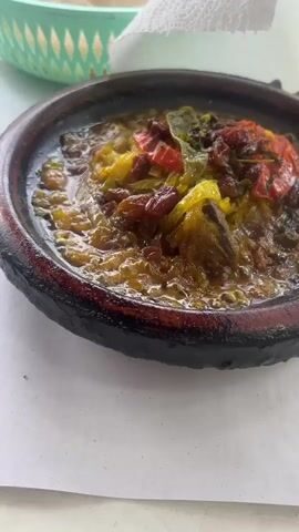 The video showcases a meal laid out on a table, likely in Boufakrane, Morocco. It opens with a close-up of a bubbling tagine dish, served in a traditional brown clay base. The tagine contains a stew of what appears to be meat, possibly chicken or lamb, with tender yellow onions, bright red bell peppers, and dark raisins, all simmering in a rich, golden sauce.

The camera then pans to reveal a side of golden-brown French fries, neatly arranged on a white plate with a blue rim. Further into the frame, three small condiment containers with colorful pink and red lids are visible, likely holding spices or sauces. A green plastic basket and a crumpled white paper towel are also on the table. The setting appears casual, with a white paper covering the table, and the meal is brightly lit, suggesting it's daytime. No people are visible, but the array of food indicates a meal prepared for consumption.
