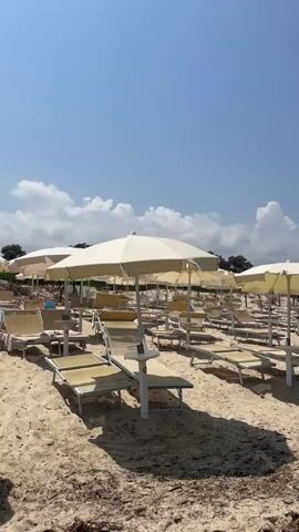This media file depicts a sandy beach with rows of empty lounge chairs and umbrellas set up. The chairs are primarily light-colored, and the umbrellas are a uniform off-white. The sand appears to be a light tan color and has some scattered debris, likely seaweed or dried vegetation, particularly in the foreground.

In the background, a small white structure with blue trim is visible, possibly a lifeguard station or a beach service hut. There are trees and greenery beyond the beach, suggesting a coastal area. The sky is blue with some scattered white clouds, indicating a sunny day, likely mid-morning to early afternoon due to the strong shadows cast by the umbrellas and chairs.

There are no people visible in the frame, suggesting the beach is either very early in the day, not yet crowded, or closed. The overall impression is one of a well-maintained but currently unoccupied beach setup, ready for visitors. The location context of Castiadas, Italy, places this scene in Sardinia, known for its beautiful beaches. The image conveys a sense of calm and anticipation, typical of a serene beach environment before the arrival of beachgoers. No visible text is present in the media file.