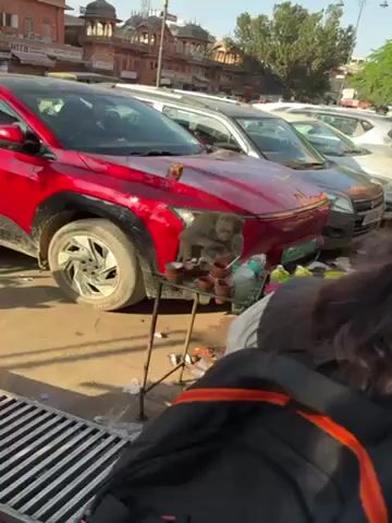 The video shows a monkey sitting on a small metal table laden with clay cups and plastic drink containers. The table is positioned in front of the hood of a red car parked alongside other vehicles. The monkey is eating from a plastic bag with a straw. A person with a black backpack walks by the camera. In the background is a tall building with a domed roof. The scene appears to be an outdoor area in India, likely a street with parked cars.