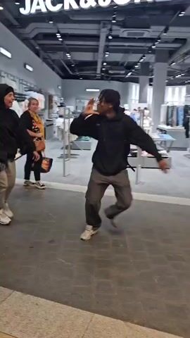 Two young males are running away from the camera in a retail environment. The first male is wearing a black hooded jacket, grey pants, and white sneakers. The second male is wearing a black jacket, grey pants, and white and grey sneakers. They are running down a walkway with stores on either side. One store visible is JACK&JONES. The walkway is tiled and there are wooden accents. The lighting is bright and appears to be from overhead fixtures.