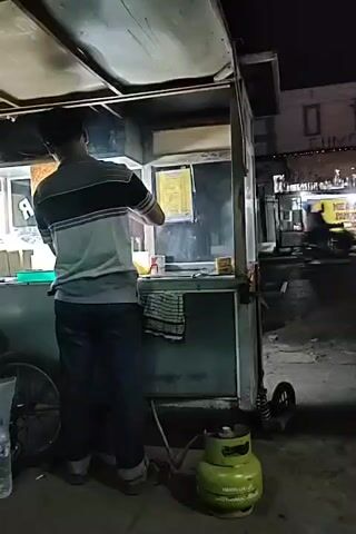 A person stands behind a food cart at night. The cart has a green propane tank attached to it. The person is wearing a striped shirt and dark pants. They appear to be preparing food, with cooking equipment visible on the cart. In the background, there are other food stalls and buildings, suggesting a busy street or market area. The lighting is dim, indicating it is nighttime.