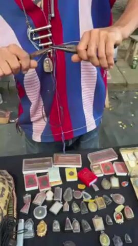 A person is shown from the torso up, standing behind a table with an assortment of small amulets or charms. The person wears a patterned short-sleeved shirt with blue, red, and white stripes and a necklace. They are using pliers to manipulate a string attached to one of the objects, likely preparing it to be added to the necklace. On the table are numerous amulets, some in small plastic cases, others laid out individually. There is also a patterned bag on the left side of the table. The setting appears to be outdoors, possibly a market or street vendor's stand.