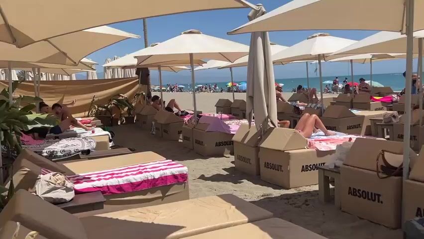 The video shows a crowded beach on a sunny day. People are lounging on beach chairs under large umbrellas. Many of the chairs are covered with cardboard boxes labeled "ABSOLUT." Some people are wearing swimwear. The beach appears to be set up for a resort or event, with lounge areas and possibly a bar or service area visible in the background. The sea is visible in the distance. People are seen walking, sitting, and interacting. There are also trash cans present on the beach.