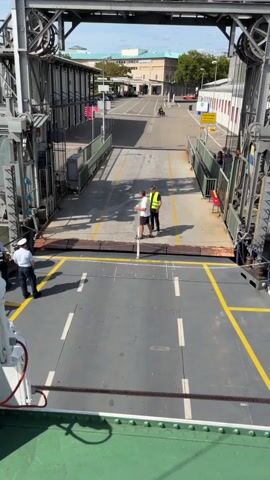 The video shows a view from a ferry, looking towards the shore. There is a ramp leading from the ferry to the shore. Two men in white shirts and dark pants, possibly crew members, are standing on the ferry's deck. One man is wearing a white hat. Two other people, one in a yellow vest and the other in a white shirt, are standing on the ramp, closer to the shore. The ramp is raised, and the gate is open. The surroundings appear to be a port or ferry terminal, with buildings and a road visible in the background. The sky is clear and blue.