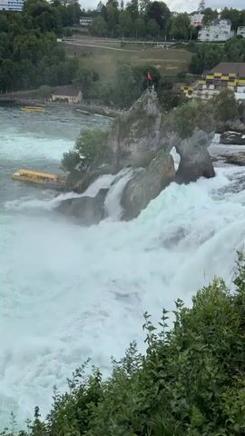 A fast-flowing waterfall with turbulent white water dominates the foreground. In the middle ground, a rocky island with a small structure and a flag on top rises from the water. A yellow boat with people is docked at the base of the island, and another yellow boat is visible further upstream. To the left, a boat with a blue top and a white canopy is moving through the choppy water. In the background, a lush green forested hillside rises, with buildings and structures visible amongst the trees. The sky is overcast. Several people are standing on a viewing platform to the left of the waterfall, observing the scene.