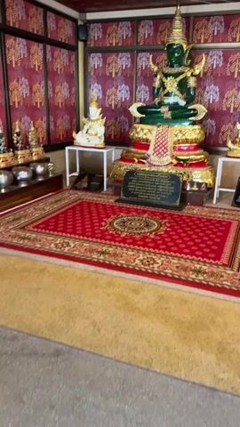 The video shows an ornate room with religious statues. In the center is a large, seated green Buddha statue adorned with gold jewelry and a tall golden headdress. The Buddha sits on a multi-tiered golden and red pedestal. In front of the Buddha statue is a dark plaque with text written in a script that appears to be Thai. To the left and right of the main statue are smaller white and gold Buddha statues, respectively, also on white pedestals. The walls are decorated with a repeating pattern of trees in shades of pink and red. A vibrant red rug with an intricate gold pattern covers the floor in front of the statues. The ceiling is paneled with wooden strips and golden trim. The overall impression is one of a richly decorated shrine or temple interior.