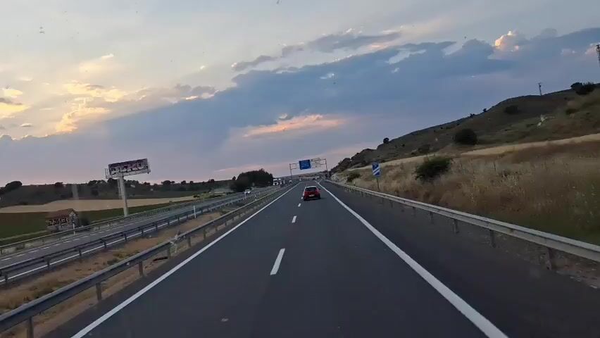 This media file captures a view from a vehicle traveling on a highway, likely in or near Medaceli, Spain. The time of day is dusk, with the setting sun casting a warm glow through scattered clouds. The sky is a mix of blues and oranges, with dramatic cloud formations.

The scene is dominated by the highway itself, with multiple lanes separated by white lines and a central barrier. Several vehicles are visible on the road, including cars and a white SUV in the foreground. The landscape alongside the highway features rolling hills, fields of dry grass, and scattered trees. In the distance, wind turbines are visible on the horizon, a common sight in rural Spain.

A key element of the footage is the side-view mirror of the vehicle, which reflects the same highway scene, offering a slightly different perspective. Within the mirror's reflection, a billboard with graffiti is visible. The overall mood is one of travel and the quiet beauty of a rural landscape at sunset. There are no people clearly visible, but the presence of vehicles and the highway indicate human activity and infrastructure.