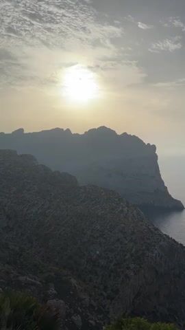 A vertical video shows a rocky coastline with the sun partially obscured by clouds in the upper left quadrant. The sun casts a hazy glow over the scene. The sea stretches out to the right, meeting the sky at the horizon. A small boat is visible in the distance on the water. In the foreground, on the right side of the frame, a palm frond is visible, suggesting a warm climate. The overall impression is a serene landscape view.