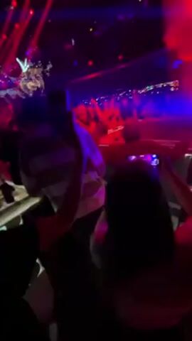 The video shows a crowded indoor club with flashing red and blue lights. People are dancing and recording with their phones. A person in a red shirt and light pants is dancing on a slightly elevated platform. There is a lit staircase to the side. A large inflatable is being bounced around in the crowd.