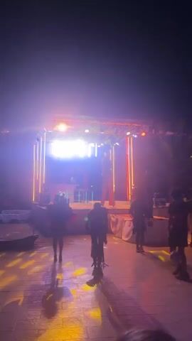 This video captures an outdoor evening event. In the foreground, several people are seated at tables, some are blurred and not clearly identifiable. In the middle ground, individuals are standing and moving. One person is visible dancing and another is walking away from the camera. There's a stage with bright lights and some vertical yellow and red striped elements. A large screen is visible in the background displaying a blue image. Flags are strung across the area. The ground appears to be a paved surface. The lighting is vibrant and colorful, with purple and yellow lights creating a lively atmosphere.