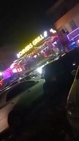 The video appears to have been taken at night, seemingly from a street-level perspective. The focus is primarily on the brightly illuminated "CHIVERS GRILL" sign, which suggests a restaurant or bar. There are multiple cars parked around the establishment. The video is a bit shaky, but you can see other sources of light in the background, possibly from other businesses or streetlights. The ground visible in the foreground appears somewhat uneven and potentially unpaved. Overall, the scene depicts a lively, nighttime environment with people likely patronizing the establishment.