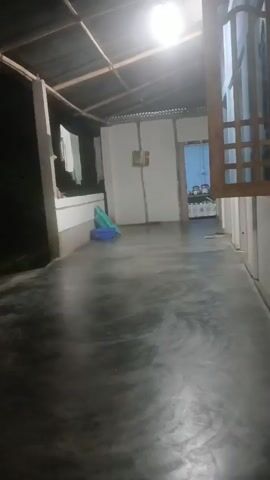 The video pans across a dimly lit, unfinished room. A single light fixture hangs from a corrugated metal ceiling, casting a bright glow. The floor appears to be concrete and is wet, with visible ripples and reflections. Two plastic containers, one blue and one green, are on the floor near a wall. There are clothes hanging on a line in the background. A doorway leads to another room, visible through a wooden frame. The interior of the second room appears to be a kitchen, with shelves and containers on display. The camera is positioned at a low angle and the footage is shaky.