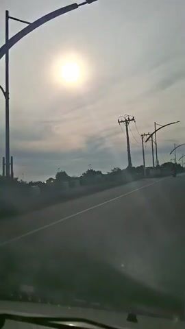 The video shows a view from inside a vehicle, looking out at a sunlit sky with scattered clouds. A road is visible in the foreground, with power lines and poles along the side. In the distance, there are trees and some buildings. The sun is bright and high in the sky, casting a glow over the scene. The vehicle appears to be moving, as suggested by the slight blur and the changing perspective of the landscape.